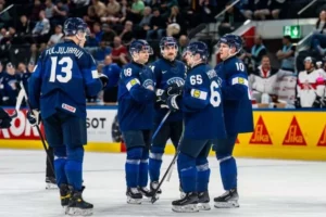 uomen maajoukkueen pelaajia sinisissä pelipaidoissa ryhmittäytyneenä jäällä ottelun katkon aikana. Kuvassa vasemmalla näkyy selin pelaaja numero 13, Jesse Puljujärvi. Keskellä ja oikealla ovat pelaajat numeroilla 18, 65 (Sakari Manninen) ja 10 keskustelemassa keskenään. Taustalla näkyy katsomo ja mainoslaitoja.