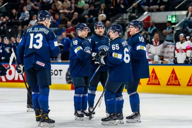 uomen maajoukkueen pelaajia sinisissä pelipaidoissa ryhmittäytyneenä jäällä ottelun katkon aikana. Kuvassa vasemmalla näkyy selin pelaaja numero 13, Jesse Puljujärvi. Keskellä ja oikealla ovat pelaajat numeroilla 18, 65 (Sakari Manninen) ja 10 keskustelemassa keskenään. Taustalla näkyy katsomo ja mainoslaitoja.