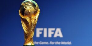 Jalkapallon virallinen kultainen MM-pokaali eli FIFA World Cup -trofee kuvattuna läheltä. Taustalla on sumennettu sininen seinä, jossa näkyy kansainvälisen jalkapalloliiton valkoinen FIFA-logo sekä sen alla teksti "For the Game. For the World.".